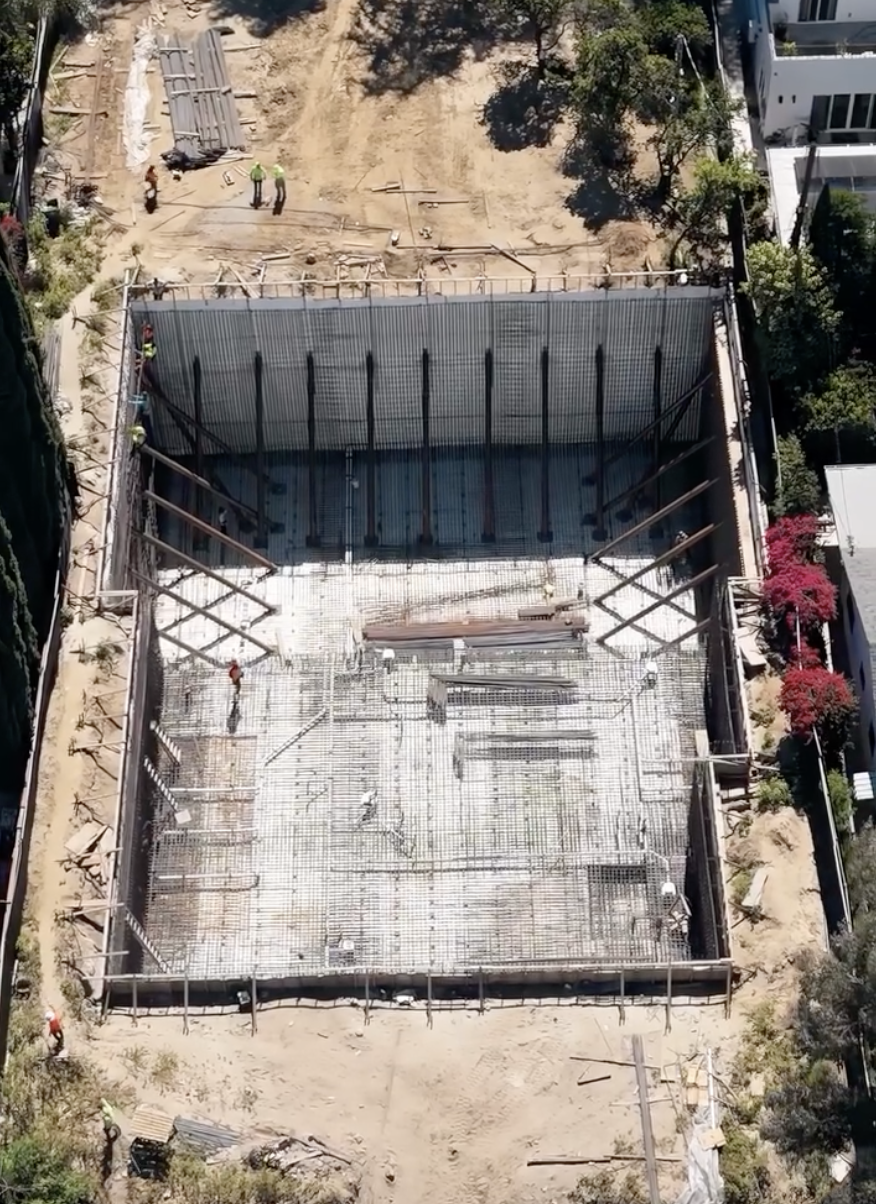 High-angle drone view of a large concrete foundation pit with steel rebar matrix and shoring walls