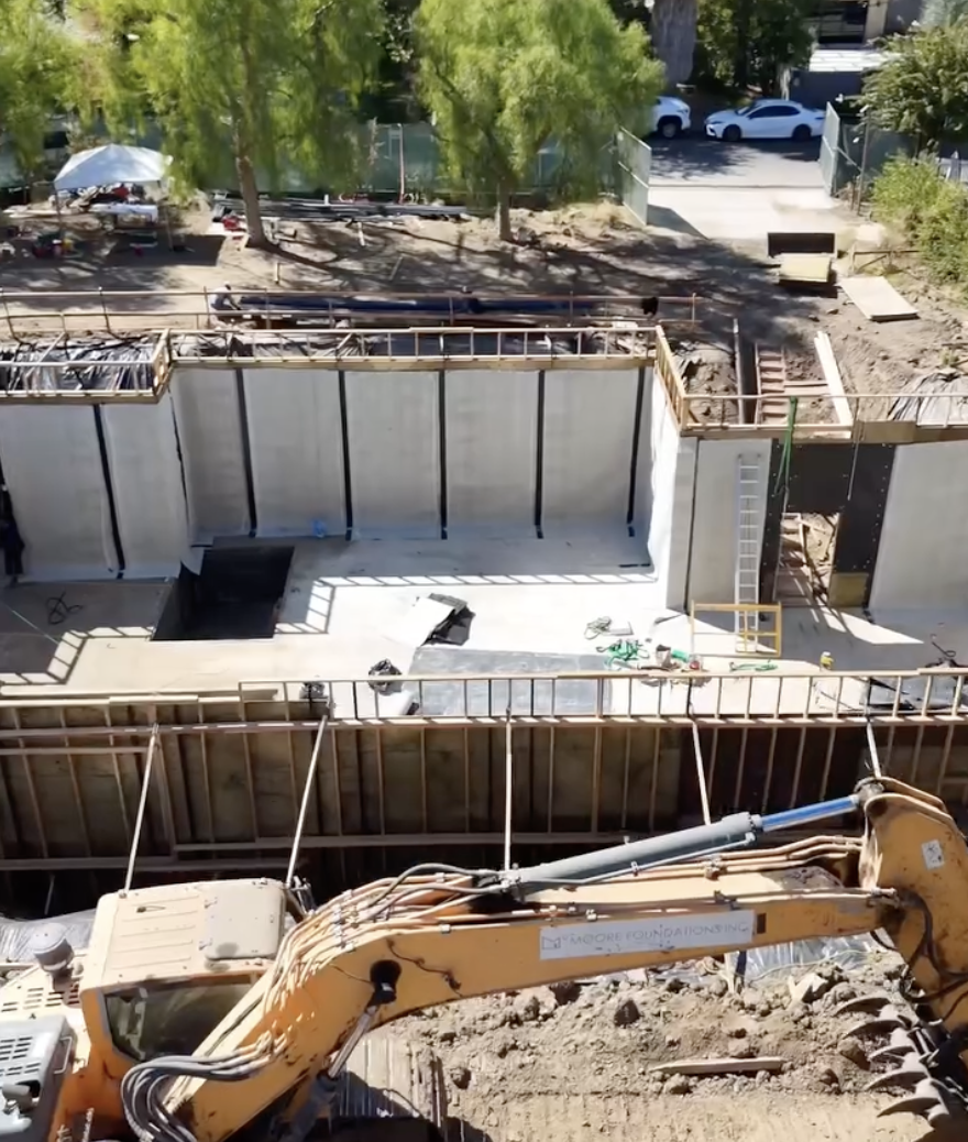 Excavator digging dirt beside concrete foundation retaining walls at a below-grade construction site