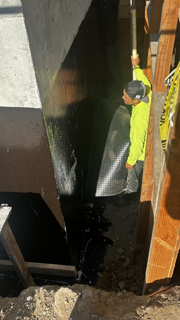 Worker installing black BITUTHENE self-adhered sheet membrane roll along foundation wall